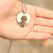 Cargar imagen en el visor de la galería, Un primer plano de una mano sosteniendo un collar con foto grabada en forma de corazón, que muestra el retrato sonriente de una mujer mayor. La mano luce un sencillo anillo en el dedo anular.