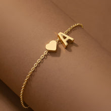 Cargar imagen en el visor de la galería, La Pulsera de tobillo con inicial es una delicada cadena de tobillo dorada con un pequeño corazón y un dije A, que se muestra en una muñeca sobre un cálido fondo marrón-perfecta para los fans de los estilos de tobillera personalizados.