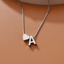 Cargar imagen en el visor de la galería, Un collar de plata con un charm en forma de corazón y un colgante con la letra A descansa sobre una superficie de color marrón claro, que combina a la perfección con la elegancia de la Pulsera de tobillo con inicial.