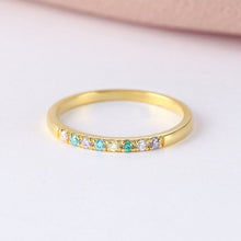Cargar imagen en el visor de la galería, Un fino Anillo de oro con piedras de nacimiento personalizado que presenta una hilera de pequeñas piedras preciosas de colores engastadas a lo largo de la parte superior, expuestas sobre una superficie blanca con un suave fondo rosa.