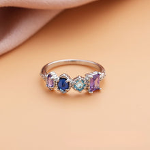 Cargar imagen en el visor de la galería, Anillo con piedras natales en plata, personalizado con cuatro piedras en tonos lila, azul oscuro, aqua y lavanda. Presentado sobre superficie durazno y fondo de tela rosa suave. Ideal como regalo para el Día de las Madres.