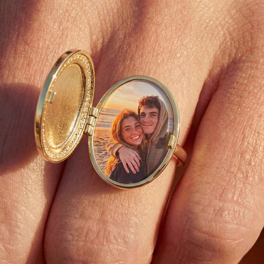 Un anillo personalizado con foto en el dedo se abre para mostrar la imagen de una pareja sonriendo al atardecer; un regalo personalizado inolvidable.