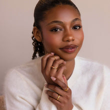 Cargar imagen en el visor de la galería, Una mujer de piel morena suave y labios brillantes lleva un jersey blanco de punto y un Anillo personalizado con piedras familiares, sonríe suavemente con las manos cerca de la barbilla sobre un fondo neutro.