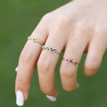 Cargar imagen en el visor de la galería, Una mano cuidada luce tres anillos Anillo personalizado Lazos del corazón -oro, plata y oro rosa-, cada uno con una pequeña piedra de nacimiento engastada sobre un fondo verde difuminado.
