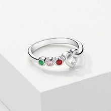 Cargar imagen en el visor de la galería, Anillo personalizado Lazos del corazón en color plateado, con tres piedras redondas verdes, rosas y rojas representando piedras de nacimiento y diseño de nudo al lado, expuesto sobre una superficie blanca geométrica.