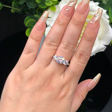 Cargar imagen en el visor de la galería, Una mano con uñas largas, doradas y brillantes luce el Anillo Tres destellos de amor en el dedo anular, sus tres piedras centelleantes brillan junto a una flor blanca y hojas verdes de fondo.