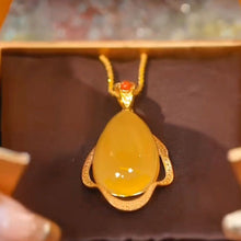 Cargar imagen en el visor de la galería, Un primer plano del Collar amuleto de piedra ágata amarilla: un colgante de ágata amarilla en forma de lágrima enmarcado en oro con una pequeña piedra roja encima, en una cadena de oro, presentado en un joyero marrón.
