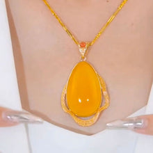 Cargar imagen en el visor de la galería, Un primer plano del Collar amuleto de piedra ágata amarilla, un collar retro de oro con un gran colgante ovalado liso de ágata amarilla y detalles dorados. Dos manos con largas uñas plateadas realzan su energía positiva sobre una prenda blanca.