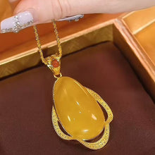 Cargar imagen en el visor de la galería, Una mano cuidada sostiene el Collar amuleto de piedra ágata amarilla, un collar retro con una gran piedra de ágata amarilla en forma de lágrima, que irradia energía positiva sobre un fondo de tela marrón oscuro.