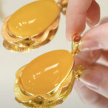 Cargar imagen en el visor de la galería, Un primer plano muestra una mano que sostiene el Collar amuleto de piedra ágata amarilla, con una gran gema amarilla en un ornamentado engaste de oro. Al fondo aparece una pieza a juego, ambas irradiando energía positiva.