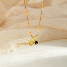Cargar imagen en el visor de la galería, Un "Collar con inicial y foto" de oro con un colgante redondo con la letra E grabada y un pequeño colgante de piedra negra, sobre tela beige, ideal para quienes buscan collares personalizados con toques elegantes.