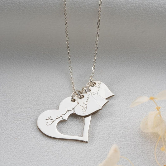 El collar Corazones de familia es un collar de plata con tres colgantes en forma de corazón de distintos tamaños, cada uno de ellos grabado con los nombres Sophia, Avery y Olivia en cursiva, sobre un fondo de tela gris claro.
