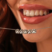 Cargar imagen en el visor de la galería, Una mujer sonriente sostiene en la boca un collar de huellitas de mascotas personalizado con cinco colgantes plateados en forma de pata, cada uno de ellos grabado con un nombre: Bentley, Franz, Findus, Mia y Minka.