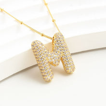 Cargar imagen en el visor de la galería, El Collar Inicial Bubble ofrece un colgante dorado con una gran letra M adornada con brillantes, exhibido sobre una superficie blanca curva. ¡Aprovecha la promo: llévate 2 por el precio de 1 y haz un regalo personalizado!.
