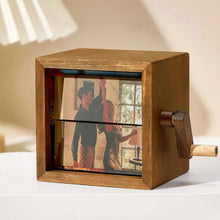 Cargar imagen en el visor de la galería, El marco fotográfico con manivela muestra una imagen giratoria de dos personas bailando junto a una ventana con un árbol al fondo. El marco descansa sobre una superficie blanca cerca de una lámpara.