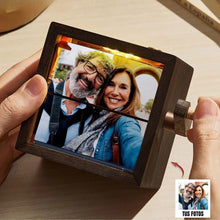 Cargar imagen en el visor de la galería, Una persona sostiene un marco fotográfico con manivela, mostrando una foto de una pareja mayor sonriente en un selfie. Un recuadro en la esquina exhibe la foto original titulada "Tus Fotos".