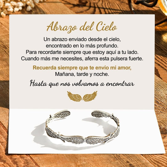 Una tarjeta con un mensaje en español sobre un abrazo del cielo se abre sobre madera, acompañada de la Pulsera Alas de ángel -símbolo de protección. La tarjeta muestra alas doradas y palabras reconfortantes, lo que la convierte en un regalo Abrazo del Cielo ideal.