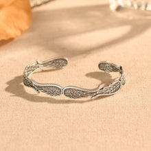 Cargar imagen en el visor de la galería, La Pulsera Alas de ángel es una pulsera de plata con detallados diseños de alas emplumadas, mostrada sobre tela beige con suave luz natural y fondo difuminado.
