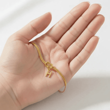 Cargar imagen en el visor de la galería, Una mano sostiene la Pulsera de nudo con inicial, con cadena dorada y colgante en forma de inicial, apoyada en la palma sobre un fondo suave y claro.