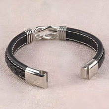 Cargar imagen en el visor de la galería, La Pulsera con nudo infinito - A mi hijo es una pulsera de piel negra con detalles plateados, diseño trenzado y cierre en forma de infinito, perfecta como regalo para hijo y que se muestra en una superficie de textura ligera.