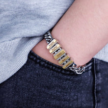 Cargar imagen en el visor de la galería, Una persona lleva una Pulsera de cadena personalizada -una pulsera de cadena de plata con dijes rectangulares de oro grabados con nombres- mientras apoya la mano en el bolsillo de unos vaqueros oscuros.
