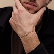 Cargar imagen en el visor de la galería, Un hombre con vello facial claro apoya la barbilla en la mano y lleva una pulsera de cubo personalizada de color dorado, un elegante regalo del Día del Padre. Viste chaqueta oscura y camisa marrón.
