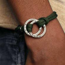 Cargar imagen en el visor de la galería, Una persona lleva una Pulsera de cuerda para hombre grabada, con dos anillos de plata entrelazados con nombres grabados, que se muestra en su muñeca cerca de unos vaqueros azul oscuro, ideal como regalo para hombre.