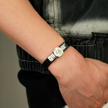 Cargar imagen en el visor de la galería, Persona que lleva en la muñeca izquierda un reloj de pulsera negro y plateado de esfera redonda, acompañado de una pulsera de cuero con nombres y foto. Visten chaqueta negra y gris y pantalón negro.