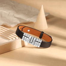 Cargar imagen en el visor de la galería, Pulsera de cuero personalizada con nombres en color negro, charms rectangulares plateados grabados, sobre superficie beige con bloques de madera y decoración; un regalo único para papá.