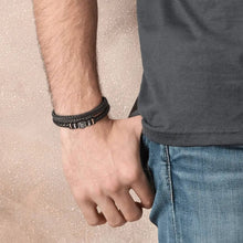 Cargar imagen en el visor de la galería, Una persona con camiseta oscura y vaqueros azules, con la mano izquierda en el bolsillo, luce en la muñeca la Pulsera de cuero - Siempre contigo Papá, una elegante pulsera de cuero trenzado negro, sobre un fondo de textura beige.