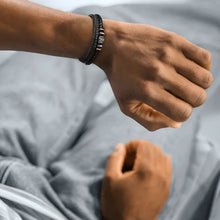 Cargar imagen en el visor de la galería, Un primer plano muestra una muñeca que lleva la Pulsera de cuero - Siempre contigo Papá, una pulsera de cuero negro con cuentas y detalles metálicos. Vestido con pantalones gris claro, su otra mano descansa sobre su regazo: perfecto como regalo para padres.