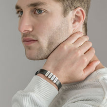 Cargar imagen en el visor de la galería, Un hombre de pelo castaño claro y ojos azules mira a un lado, apoyando la mano en el cuello. Lleva un jersey claro y una pulsera de hombre con nombres grabados con varios pequeños dijes rectangulares plateados.