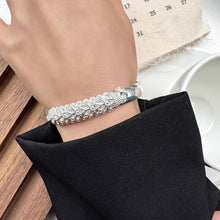 Cargar imagen en el visor de la galería, Una mano adornada con la Pulsera Elegancia Real, con un elegante motivo de hojas en acero inoxidable, está parcialmente cubierta por una funda negra, con un calendario y una copa blanca visibles en el fondo.