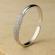 Cargar imagen en el visor de la galería, La Pulsera Elegancia Real es un brazalete de plata con un intrincado motivo de pavo real y plumas detalladas, lucido sobre una superficie de piedra beige.