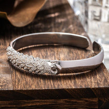 Cargar imagen en el visor de la galería, La Pulsera Elegancia Real es una pulsera de plata acero inoxidable con un diseño ornamentado en forma de hoja y pequeñas piedras incrustadas, elegantemente expuestas sobre una superficie de madera oscura.