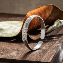 Cargar imagen en el visor de la galería, La Pulsera Elegancia Real, una pulsera de plata con detallados diseños florales y de hojas, se alza sobre una superficie de madera con hojas secas de fondo.
