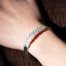 Cargar imagen en el visor de la galería, Un primer plano de la Pulsera Elegancia Real, con un intrincado diseño en forma de pluma de acero inoxidable en la muñeca de una persona, sobre un fondo marrón texturizado.