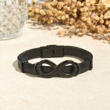 Cargar imagen en el visor de la galería, La Pulsera Infinito - A mi Hombre, una elegante pulsera de cuero negro con un charm del símbolo del infinito, se muestra en una superficie clara junto a flores secas y cristal texturizado: un regalo ideal para hombre.