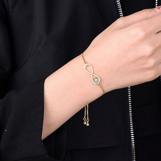 Una mano lleva la Pulsera Infinito con ojo turco (OFERTA 2X1), con un símbolo dorado del infinito y un amuleto de protección contra la energía negativa, sobre una prenda negra con cremallera.