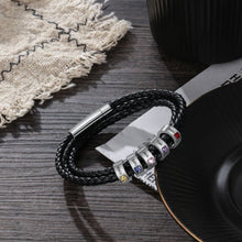 Cargar imagen en el visor de la galería, La Pulsera Piedras de vida - A mi Padre, una pulsera de cuero trenzado negro con dijes de plata grabados con nombres y piedras de nacimiento, se coloca en un plato negro junto a una tela tejida: un regalo ideal para el Día del Padre.
