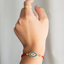 Cargar imagen en el visor de la galería, Una mano con un anillo de oro y la Pulsera protectora de ojo turco -una pulsera roja con un amuleto de ojo con piedras azules y blancas- destaca sobre un fondo suavemente difuminado.