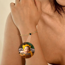Cargar imagen en el visor de la galería, Primer plano de la muñeca de una mujer que lleva la Pulsera Recuerdo eterno, una delicada pulsera de oro con un colgante circular negro. En el recuadro, un bebé dormido con un gorrito amarillo, lo que lo convierte en un regalo personalizado ideal.