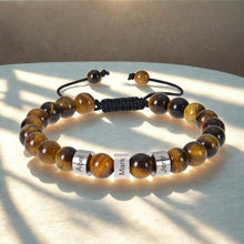 Cargar imagen en el visor de la galería, A Pulsera tejida personalizada con nombres, hecha con cuentas de ojo de tigre marrón y dorado pulido y tres abalorios plateados grabados con Abel, Mark y Jerry, es una pulsera especial para papá exhibida bajo la luz del sol.