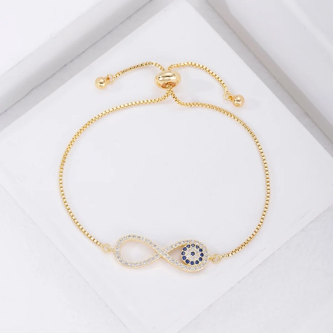 La Pulsera Infinito con ojo turco (OFERTA 2X1) es una pulsera ajustable de oro con un símbolo de infinito y piedras azules, inspirada en la tradicional pulsera de ojo turco para la protección contra la energía negativa, que se muestra sobre un fondo blanco.