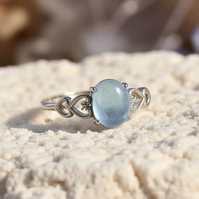 El Anillo ajustable Cristal de Mar presenta una pulsera de plata con filigrana en forma de corazón y un cristal de mar ovalado azul pálido en el centro, que se muestra sobre una superficie blanca texturizada.