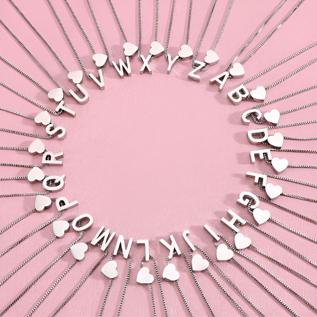 Los collares de plata con colgantes en forma de corazón e iniciales de la A a la Z están dispuestos en círculo sobre un fondo rosa, cada uno con una letra y un charm, ideales para combinar con la Pulsera de tobillo con inicial para formar un conjunto a juego.