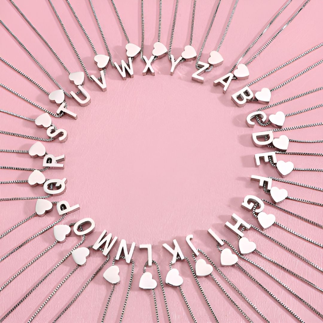 Los collares de plata con colgantes en forma de corazón e iniciales de la A a la Z están dispuestos en círculo sobre un fondo rosa, cada uno con una letra y un charm, ideales para combinar con la Pulsera de tobillo con inicial para formar un conjunto a juego.