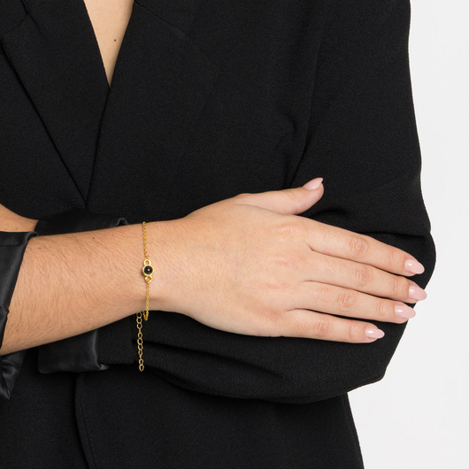 Una persona con americana negra cruza los brazos, mostrando la Pulsera con foto, una pulsera de oro personalizada con un pequeño colgante negro y dorado. Lleva las uñas cuidadas en un tono suave y natural.