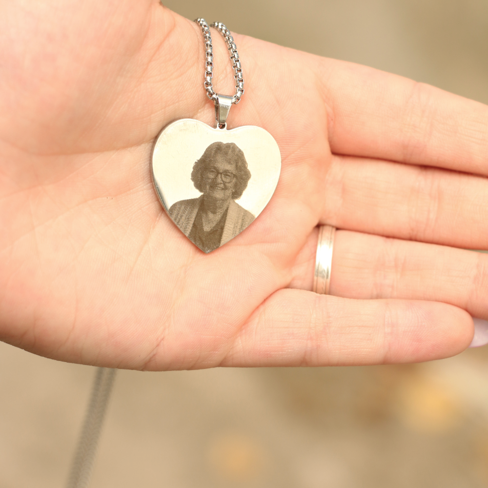 Una mano sostiene un Collar con Foto Grabada en Forma de Corazón con una foto grabada de una mujer mayor con gafas y rebeca. La persona lleva un sencillo anillo en el dedo.
