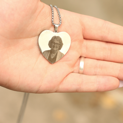 Una mano sostiene un Collar con Foto Grabada en Forma de Corazón con una foto grabada de una mujer mayor con gafas y rebeca. La persona lleva un sencillo anillo en el dedo.